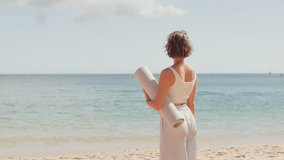 Woman holding yoga mat and smiling while turning towards the camera on sandy beach, enjoying peaceful ocean view, representing positivity and calmness - Powered by Shutterstock - Get 15% off with code: PIKWIZARD15