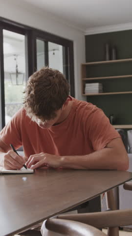 Vertical video: Man writing in notebook near glass doors tapping pen gazing before resuming writing. Thoughtfulness, concentration, creativity, workspace, reflection, learning, vertical video