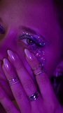 Close-up of a young woman gently touching her cheek with glitter makeup under violet neon light. Futuristic alien vibe, soft chrome nails, dreamy and mysterious expression - Powered by Shutterstock - Get 15% off with code: PIKWIZARD15