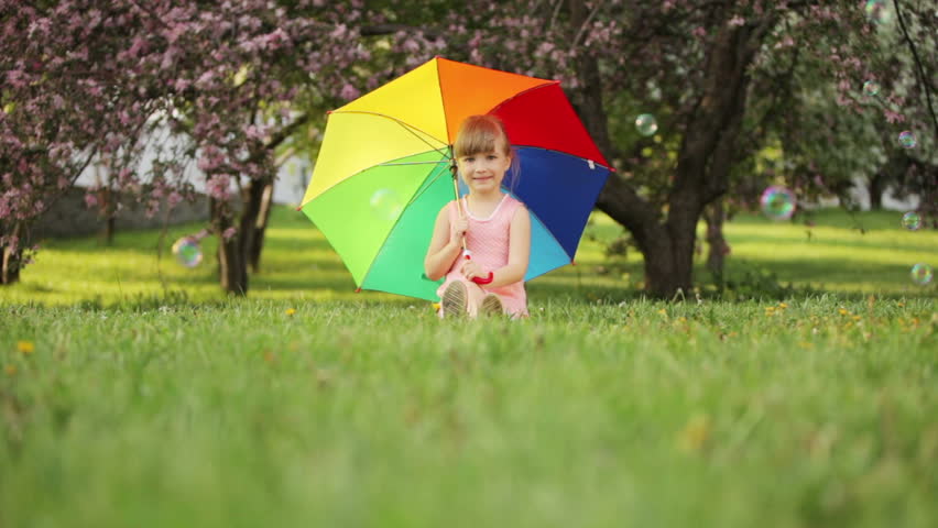 Little girl sitting in garden with umbrella and smiling
