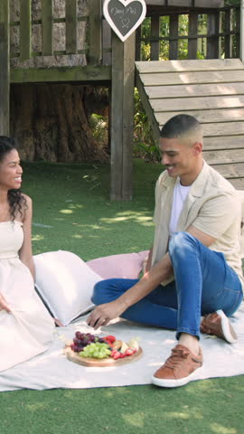 Vertical video: Diverse couple pulling smartphone, snapping selfies in backyard with Marry Me sign. Romance, picnic, leisure, rustic, happiness, outdoor, vertical video