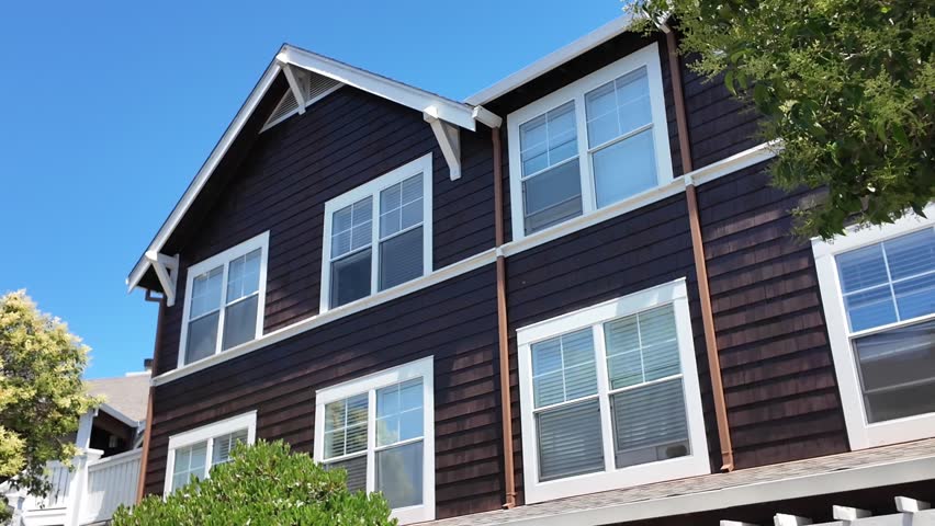 A large brown house with many windows and a slanted roof. The house is surrounded by trees and has a blue sky in the background
