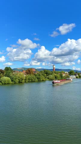 A barge glides smoothly along a tranquil river surrounded by lush greenery and a picturesque backdrop of hills and clouds on a sunny afternoon, vertical video