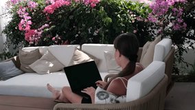 Back view captures woman on sofa facing flowers, doing remote work in garden lounge. Work rhythm stays relaxed yet productive, work framed by pink blooms and natural light - Powered by Shutterstock - Get 15% off with code: PIKWIZARD15