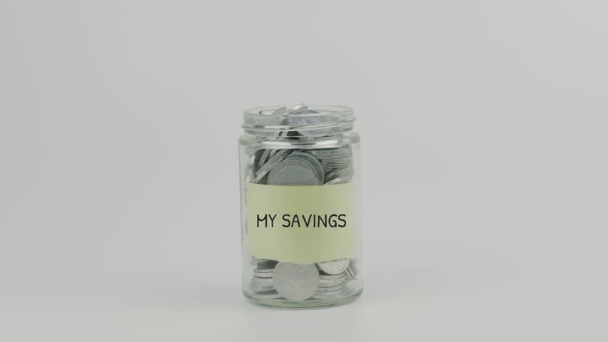My Savings word text on yellow sticky note attached to coin jar, hand inserting coin and sealing lid on white background, symbolizing personal savings, financial planning, and wealth building