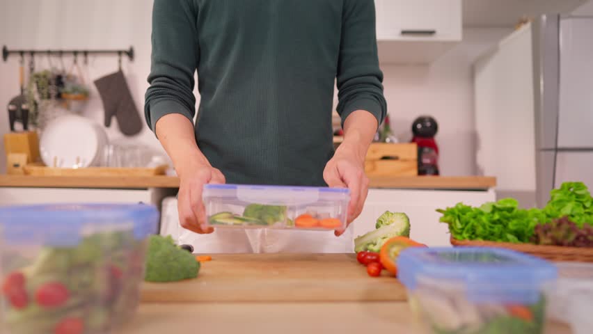 Close up of man cooking healthy foods in kitchen in morning at home. Attractive male feeling happy and relax, enjoy preparing ingredients and making green salad for breakfast in lunch box in house.