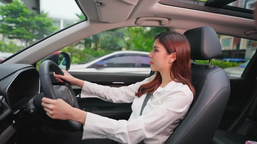 Beautiful Asian woman fastening her seatbelt while driving a car. Attractve young woman employee sit inside the vehicle, feel happy and enjoy showing safe and protected driving while driving to work.