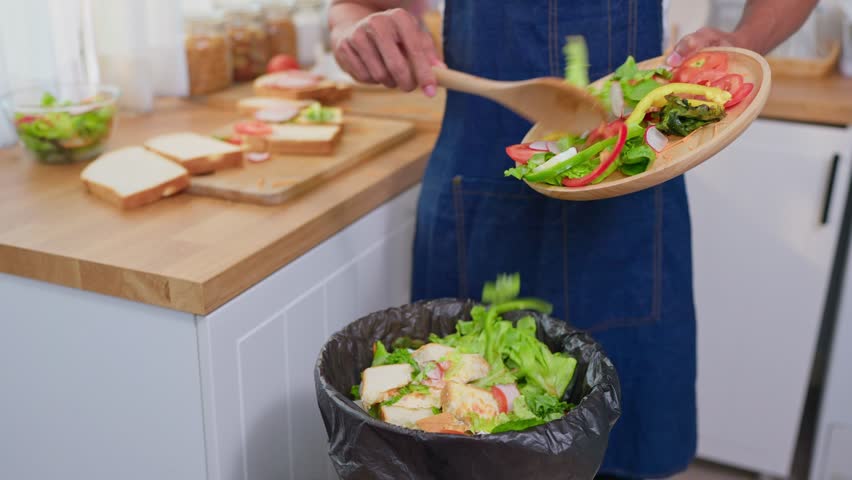Close up of man in kitchen apron throwing away fresh food scraps into trash bin. The bin is filled with vegetables and salad leftovers, showing household food waste and kitchen disposal in house.
