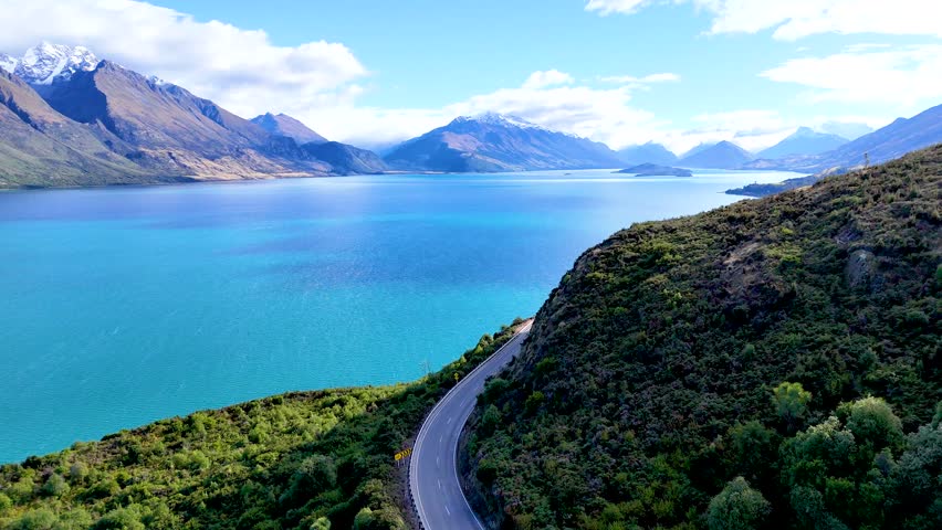Drone glides above winding road, turquoise lake, and mountains under bright daylight in Queenstown