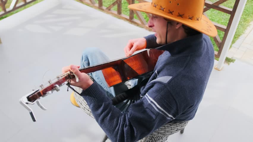 Young male musician in a cowboy hat performs on acoustic guitar while singing during an outdoor music video shoot. Emotional expression, cinematic scene rooted in folk and country aesthetics.