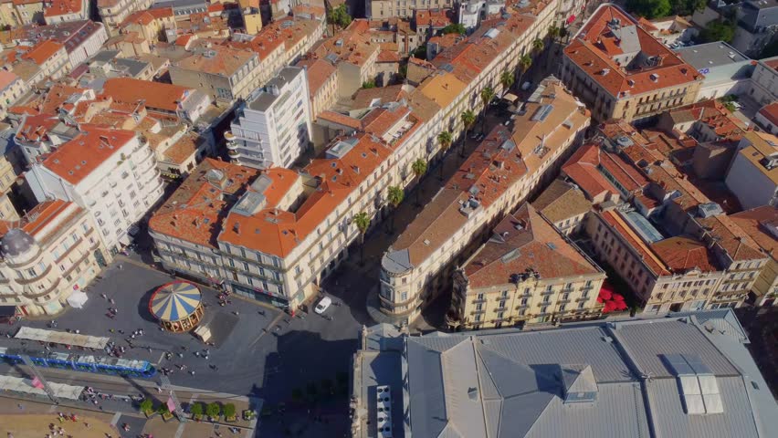 An aerial drone shot captures the vibrant and historic city center of Montpellier, France, showcasing its charming architecture, terracotta rooftops, and bustling public spaces, including a carousel i