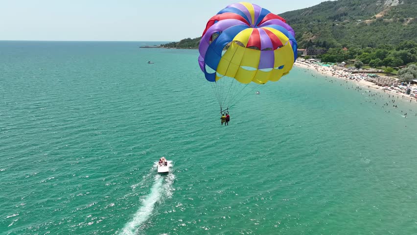 Speedboat pulling a parachute with people moving behind the boat over the water. Aerial view of a speedboat pulling a parachute against a seaside resort. Summer vacation photo.