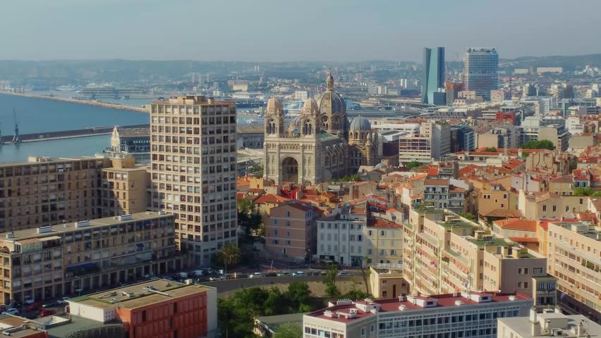 Aerial footage capturing the iconic cityscape of Marseille, France, featuring the prominent Notre-Dame de la Garde Basilica overlooking the vibrant city, the bustling Old Port, and modern architecture