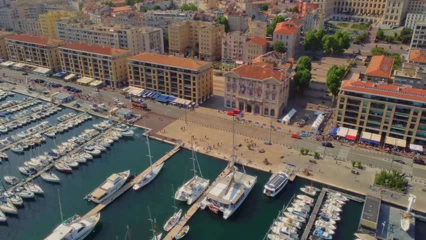 A drone footage capturing the vibrant Old Port (Vieux-Port) of Marseille, France, showcasing numerous boats docked in the marina alongside the picturesque historic buildings and bustling promenade.