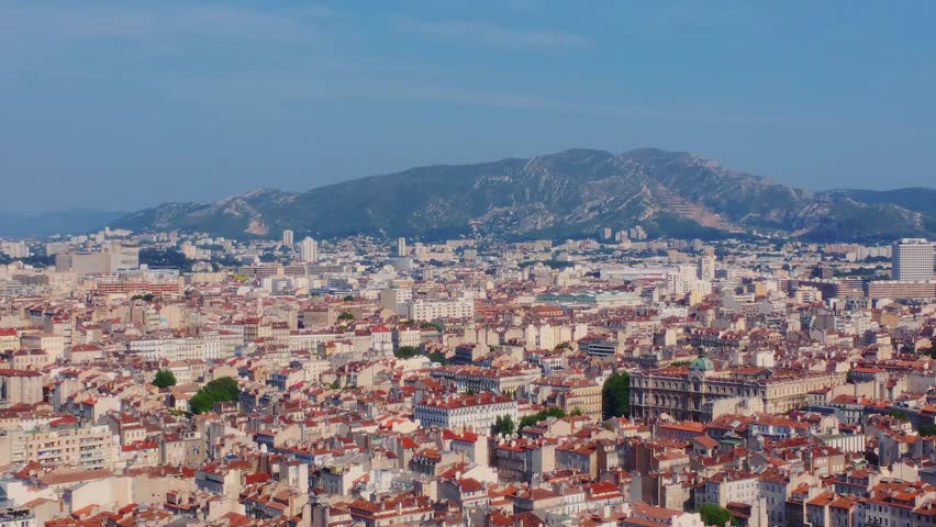 aerial view captures the vibrant cityscape of Marseille, France, under a clear blue sky, featuring the iconic Notre-Dame de la Garde Basilica prominently overlooking the terracotta rooftops and dense