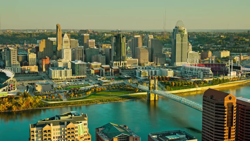 A retreating aerial of downtown Cincinnati from Covington, Kentucky in the morning, showcasing the skyline, Ohio River, and Roebling Bridge under golden sunrise light. Perfect for travel and urban.