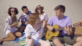 Group of friends relaxing on the beach while a young man plays guitar. - Powered by Shutterstock - Get 15% off with code: PIKWIZARD15