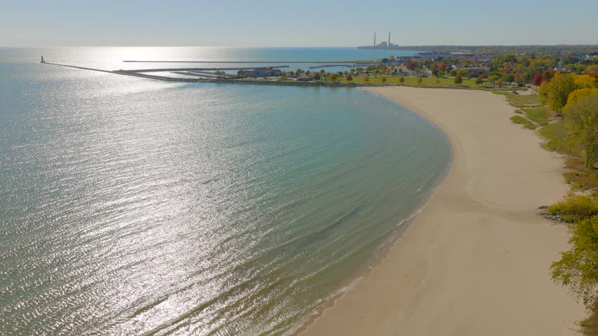 Drone flyover of Lake Michigan shoreline in Sheboygan, Wisconsin toward the harbor with sparkling sunlight on the water and colorful autumn scenery.