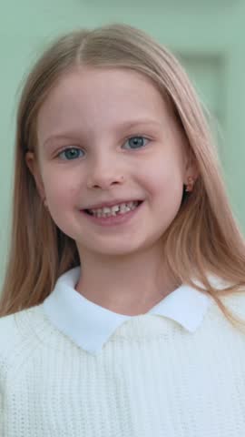 Blond-haired girl with blue eyes smiling wearing white sweater expressing genuine happiness and joy. Girl kid child orphan in orphanage. Vertical video