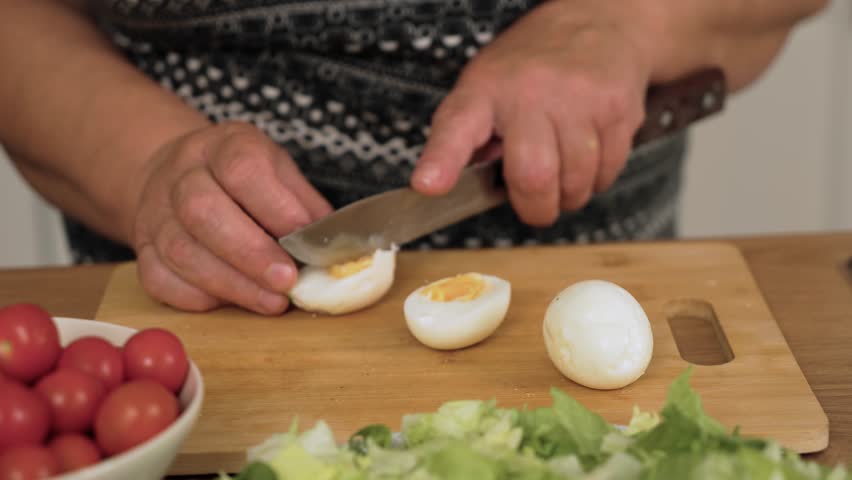 Cutting boiled egg for preparing Caesar salad. Slicing hard cooked egg for making fresh dish. Halving protein ingredient for Caesar recipe. Preparing egg pieces for vegetable mix