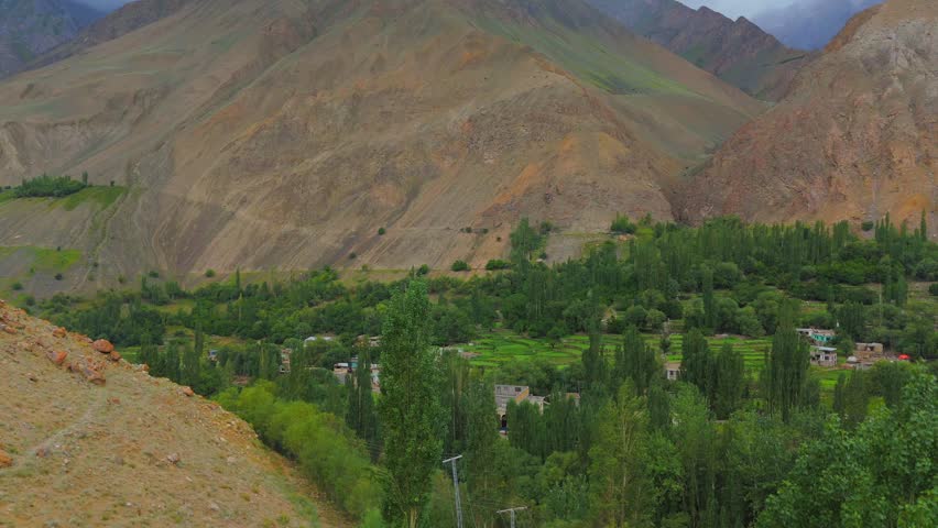 Aerial view of a vibrant green valley with cultivated fields nestled between rugged mountains under a cloudy sky, Thally, Gilgit Baltistan, Pakistan.