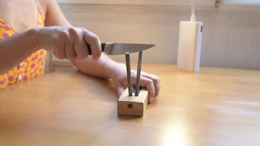 Close-up of a person sharpening a kitchen knife with a manual knife sharpener on a wooden table. Concept of cooking, preparation, kitchen tools, safety, and home maintenance
