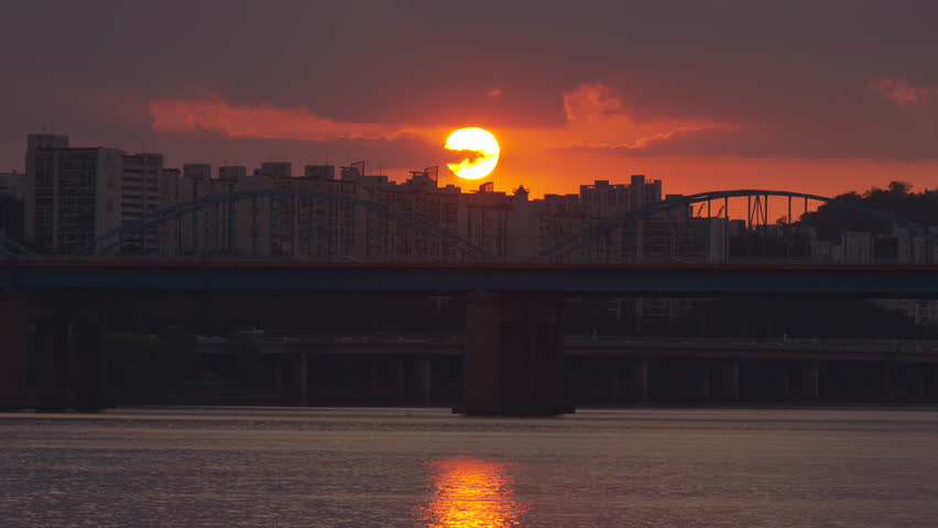 Sunset over Seoul’s Dongjak Bridge with the Han River glowing below. High-rise apartments silhouette against the fiery sky as the sun dips, blending urban landscape with natural beauty.
