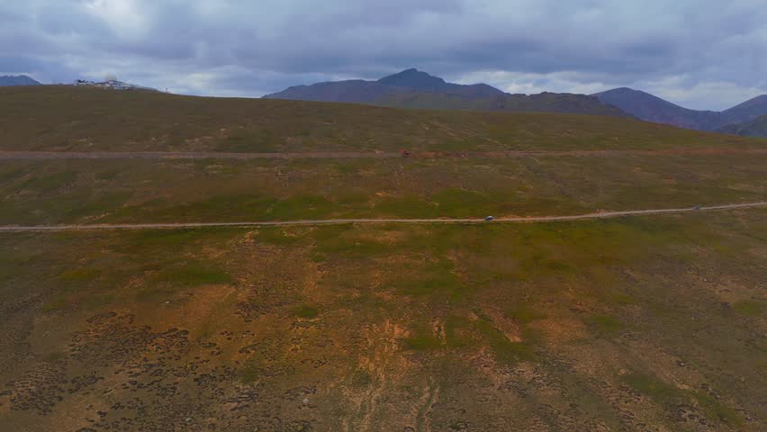 Aerial view of vehicles on Deosai Park Road, cutting through grassy hills, set against distant mountains and an overcast sky, Skardu, Gilgit Baltistan, Pakistan.