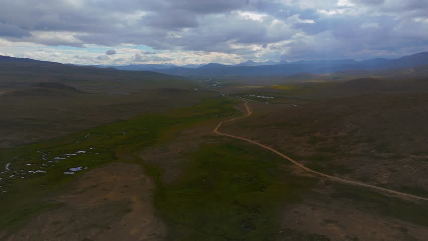 Aerial view of Deosai Park Road winding through a vast landscape, contrasting with the distant mountains and cloudy sky, Skardu, Gilgit Baltistan, Pakistan.