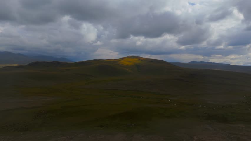 Aerial view of a vast, grassy landscape with rolling hills, illuminated by a golden light piercing through the dramatic, cloud-filled sky, Skardu, Gilgit Baltistan, Pakistan.