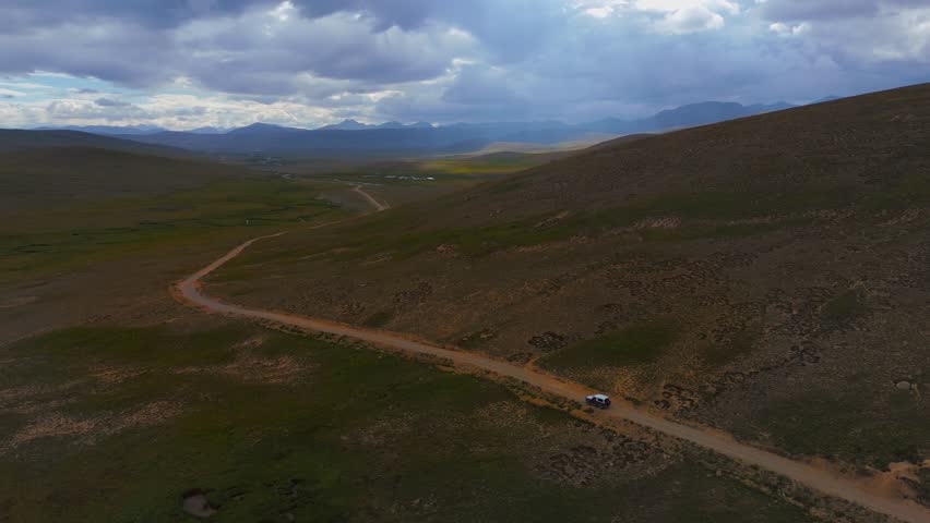Aerial view of a lone vehicle traversing the winding Deosai Park Road through a valley, contrasting the brown hills and dramatic sky, Skardu, Gilgit Baltistan, Pakistan.