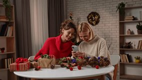 Two women in warm sweaters happily preparing handmade gifts. The friends communicate via video call on a smartphone. The decorated tree and festive decor create a cheerful atmosphere for celebration. - Powered by Shutterstock - Get 15% off with code: PIKWIZARD15