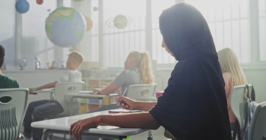 Young Muslim Girl Wearing Hijab Sitting at the Desk, Writing School Exam in Notebook, Doing Task or Drawing. In the Background, Children Studying in Bright Classroom of Modern Elementary School.