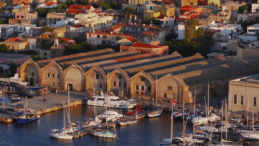 Aerial view of Chania town in Crete, Greece, highlighting a domed church, white buildings, rugged mountains, and warm golden hour lighting.