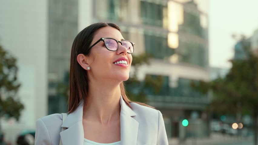 Portrait of Latin successful businesswoman in eyeglasses standing outdoor looking dreaming aside at sky. Thoughtful confident proud beautiful woman entrepreneur smiling on skyscrapers city background