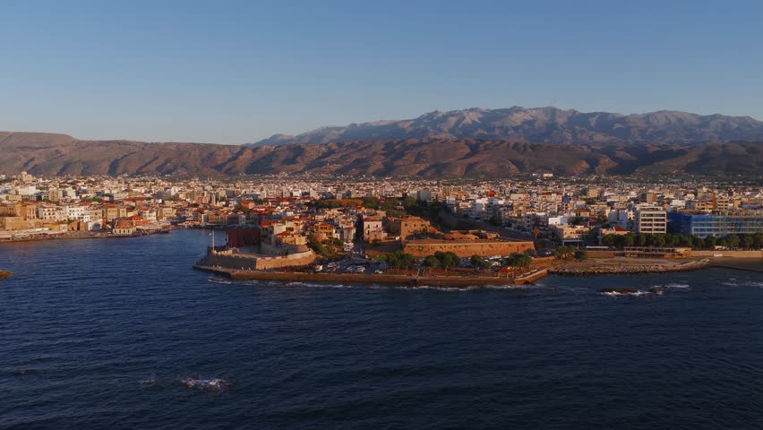 Aerial view of Chania town in Crete, Greece, with a sweeping motion over the Venetian harbor, lighthouse, and surrounding Aegean Sea at sunset.