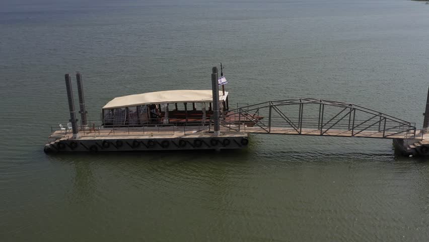 A serene view of the Sea of Galilee (Kinneret) with a floating dock and pier. The tranquil waters are surrounded by rolling hills and distant buildings, capturing the calm and natural beauty of the re