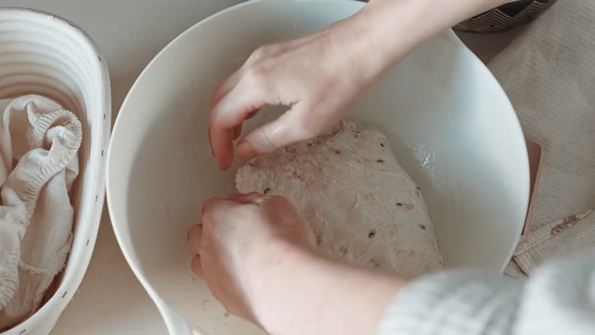 Close-up video of hands stretching and folding bread dough with seeds in a white bowl during sourdough bulk fermentation.