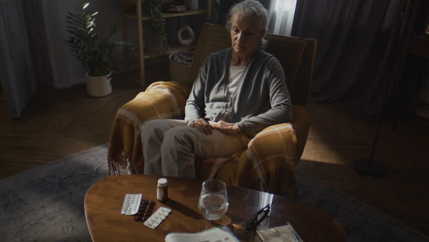 Senior woman checks pill bottle doubting of therapy plan at home. Old lady takes medication container from coffee table with glass of water in living room