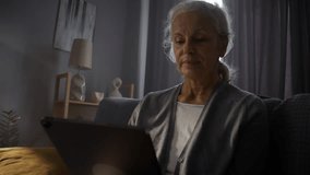 Happy elderly woman waves hand Hello to digital tablet making video call at home. Grey haired lady talks to grandchildren via mobile device in living room - Powered by Shutterstock - Get 15% off with code: PIKWIZARD15