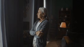 Elderly woman with crossed arms gazes pensively through window at home. Old grey-haired lady expresses solitude and quiet contemplation in shady living room - Powered by Shutterstock - Get 15% off with code: PIKWIZARD15