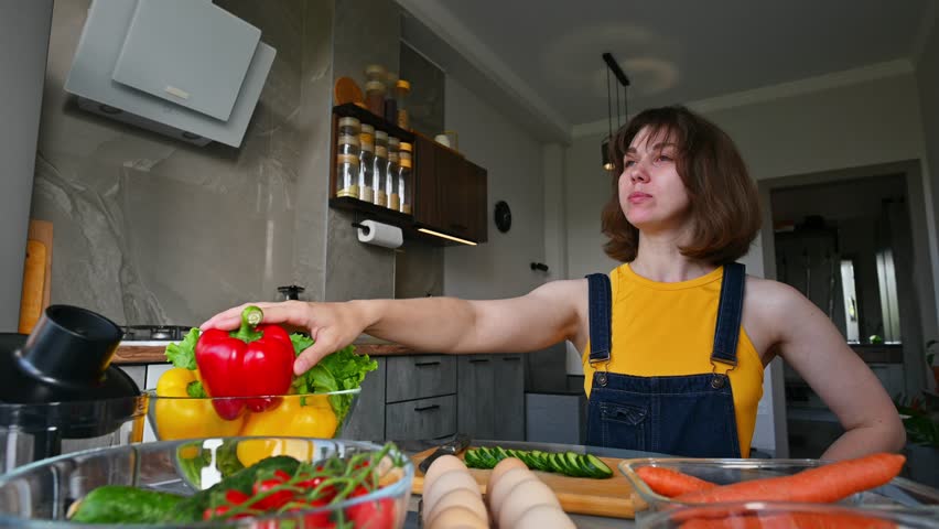 Positive young attractive Caucasian woman heroically eating freshly sliced cucumber in home kitchen surrounded by vegetables