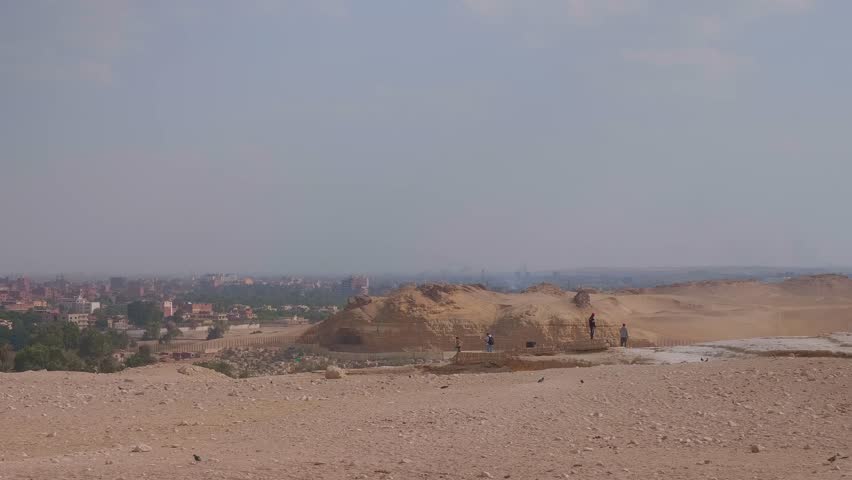 A wide shot capturing the vast desert landscape of an ancient Egyptian site, with remnants of structures or tombs in the foreground, and a modern cityscape visible in the hazy distance under a clear s