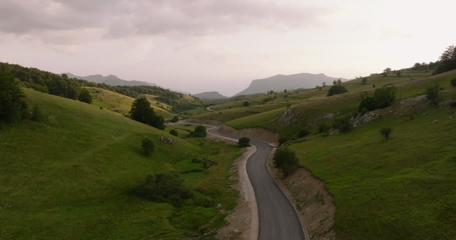 Winding road through a hilly landscape The route cuts through green hills and meadows The sky is overcast Trees dot the scenery