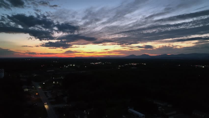 Mystic golden sunset at horizon over dark american city with deriving car on road. Aerial panorama view. Suburb city neighborhood at golden hour. Headlights of vehicle on asphalt.