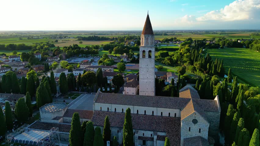 Aerial view of Basilica di Santa Maria Assunta at sunset in Aquileia, Italy, surrounded by historic ruins, charming houses, and green fields. Famous UNESCO World Heritage site with rich Roman history 