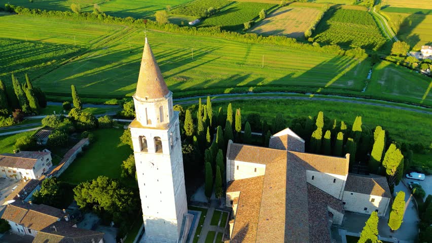 Aerial view of Basilica di Santa Maria Assunta at sunset in Aquileia, Italy, surrounded by historic ruins, charming houses, and green fields. Famous UNESCO World Heritage site with rich Roman history 