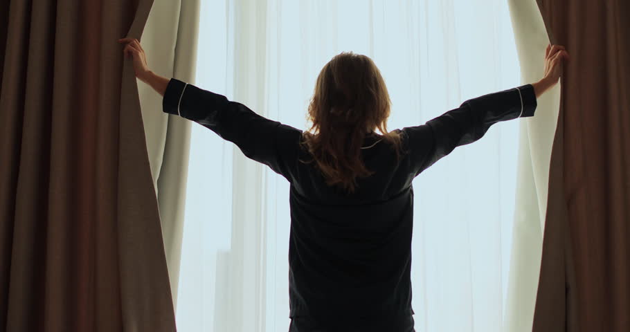 Wearing cozy pajamas, female draws back the curtains. Female hands open window curtain in morning.