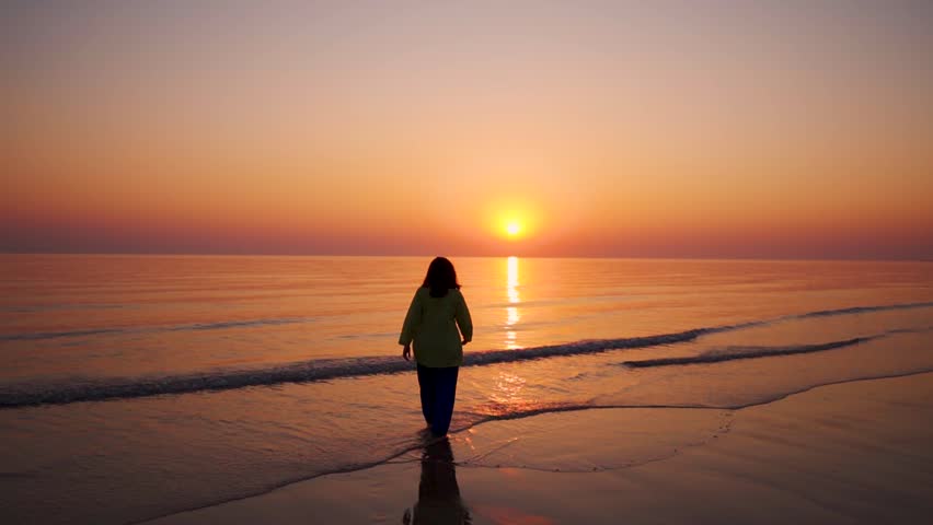 Slow motion shot of young woman walk on beach during sunset at Mandvi, Gujarat, India. Summer holidays at tropical island. Small waves crashing on shore and sun setting on horizon. Relaxation concept 