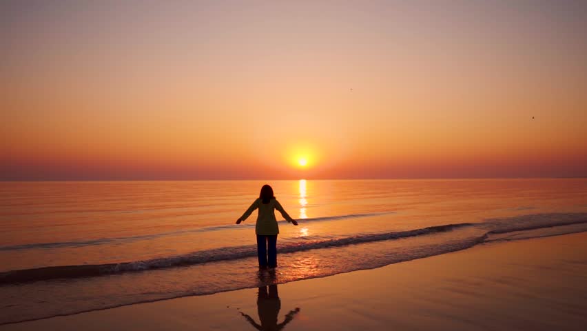 Young woman raising hands on beach during sunset at Mandvi, Gujarat, India. Summer holidays at tropical island. Small waves crashing on shore and sun setting on horizon. Relaxation concept 